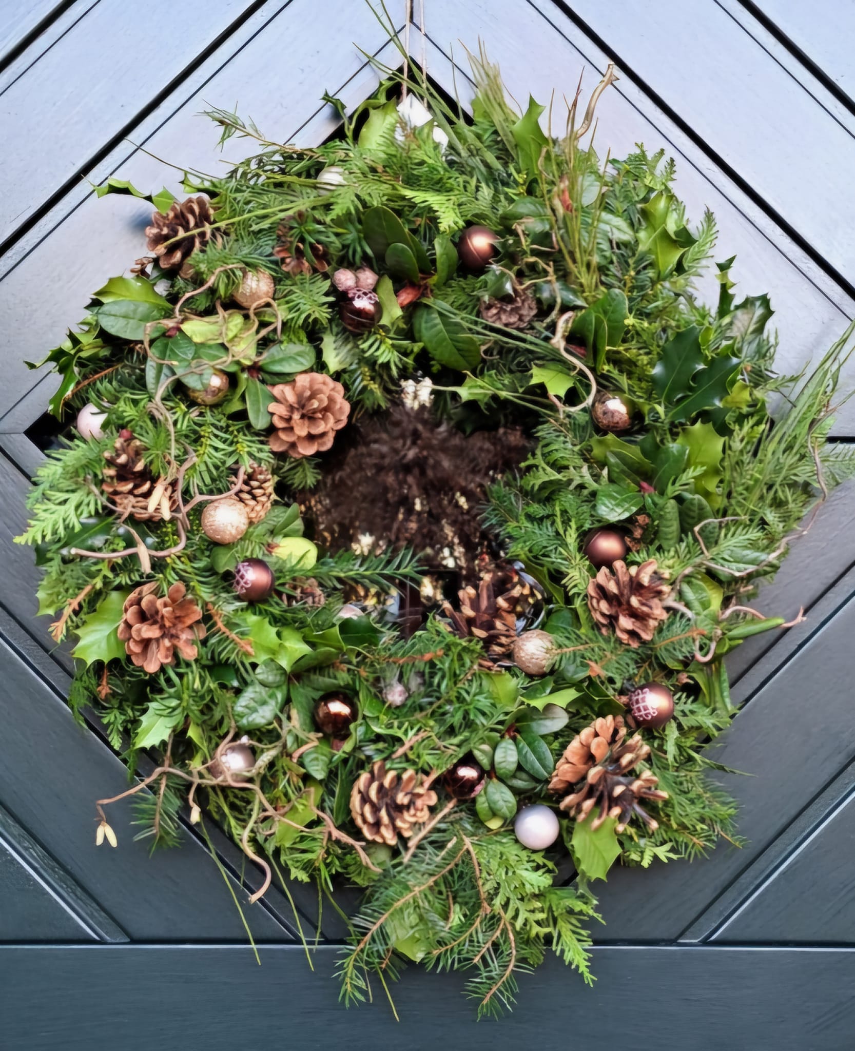 Haustürkranz aus Eibe, Ginster, Ilex, Korkenzieherhasel und Rhododendron aus dem Garten, gewickelt mit Draht auf einen großen Strohrömer, dekoriert mit Zapfen und kleoöinen Kugeln