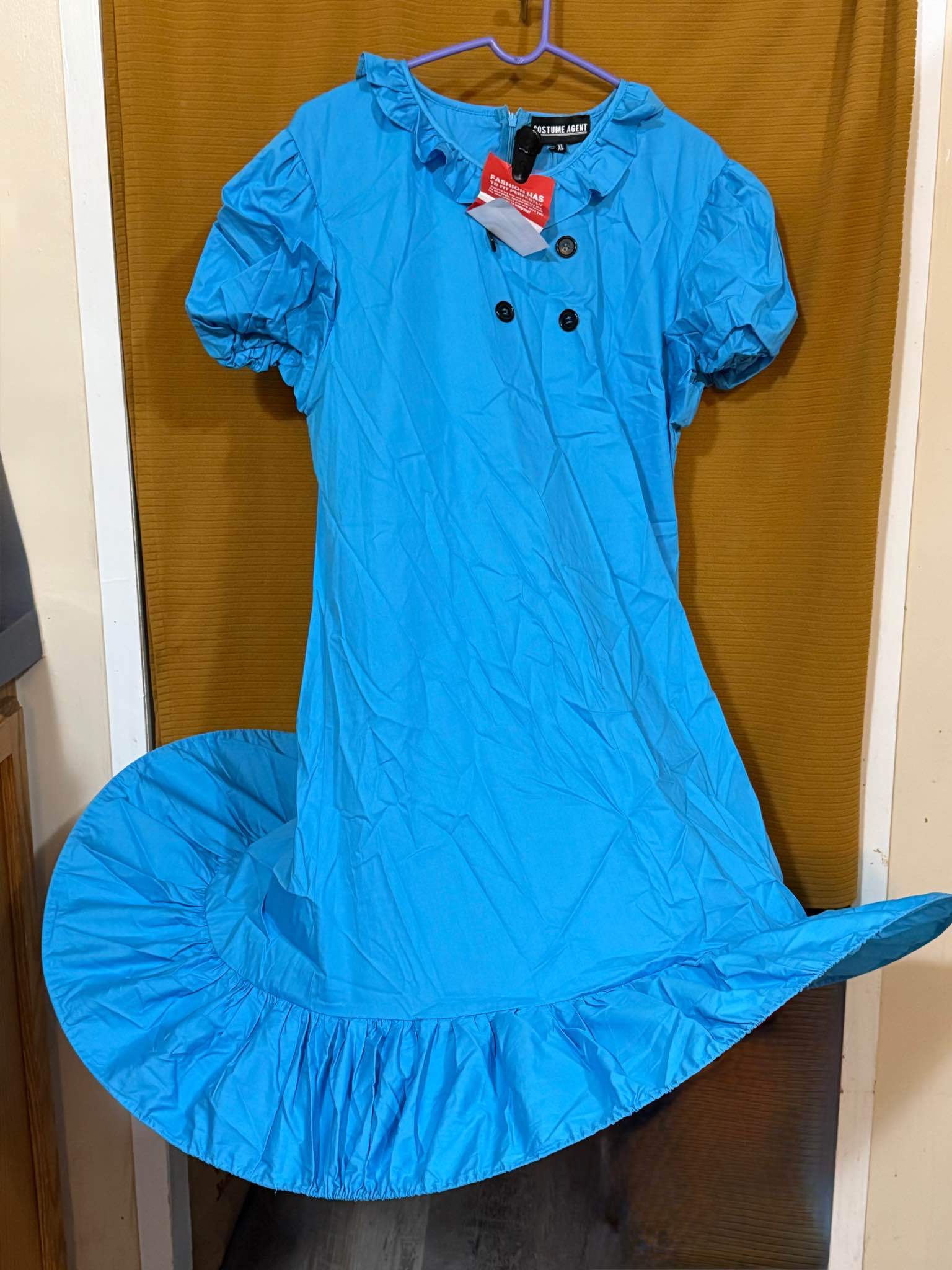New Costume Agent Lucy Dress in size XL. This blue adult costume dress has a ruffled collar, short puff sleeves, and a double-breasted button front. It is made of a lightweight fabric