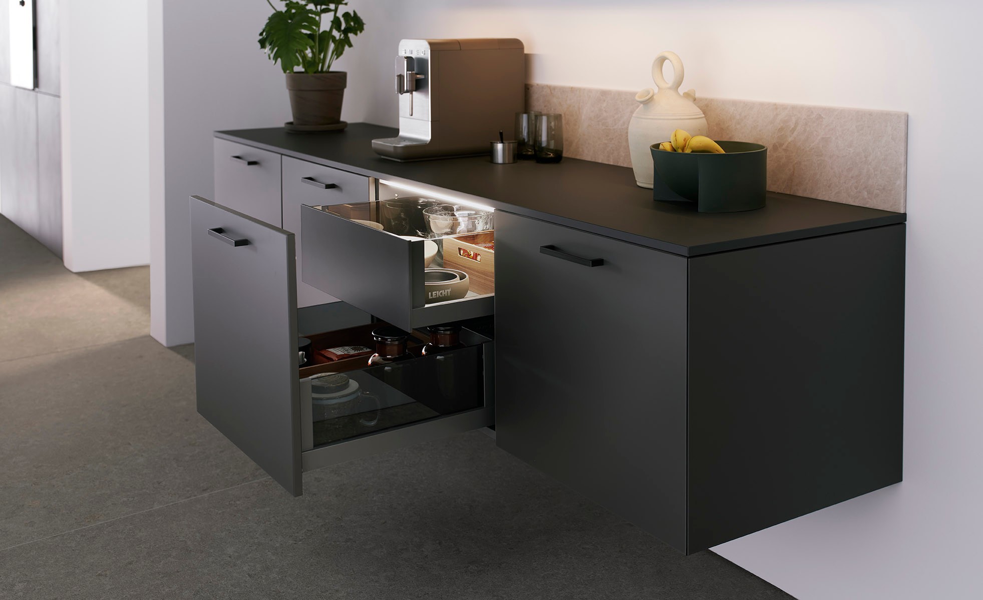 Sideboard in Carbongrau Ein Sideboard in carbongrau, das Platz für Kaffeemaschine oder andere essentielle Küchenutensilien bietet. Über dem Möbel verläuft eine auf die gesamte Breite geplante Naturstein-Leiste in ROCCA, die einen unmittelbaren