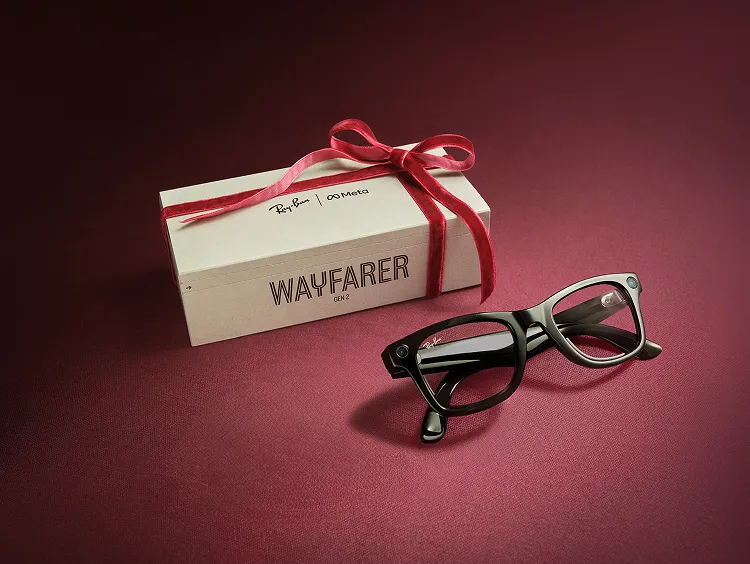 Ray-Ban Meta Wayfarer AI Glasses (Gen 2) i Shiny Black visas hopvikta på en mörkröd yta bredvid sin ursprungliga låda, som slagits in med ett rött band som en gåva.