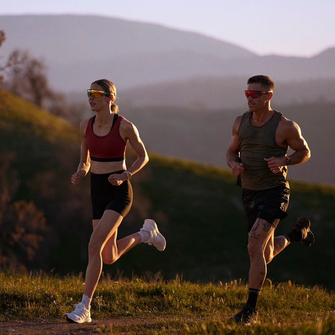 A woman and man running outdoors while both wearing Oakley Meta glasses.